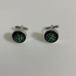 Puede incluir: Un par de gemelos plateados con diseño de brújula. Cada gemelo presenta una esfera negra con indicaciones direccionales verdes y blancas. Los gemelos tienen forma redonda clásica y un poste plateado para sujetarlos.