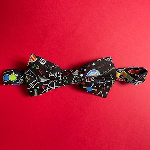 May include: A black bow tie with a science-themed print featuring colourful chemical formulas, diagrams, and symbols. The bow tie is on a red background.