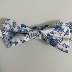 Może przedstawiać: Biała mucha z niebieskim kwiatowym wzorem. Mucha wykonana z tkaniny ma powtarzający się wzór niebieskich kwiatów i liści. Mucha jest zawiązana w klasyczny kształt kokardy.