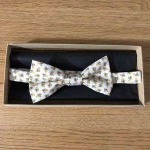 May include: A white bow tie with a pattern of yellow bees. The bow tie is in a box with black lining.