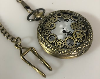 May include: A gold-toned pocket watch with a chain and a decorative clasp. The watch face has a white background with black hands and Roman numerals. The watch face is surrounded by a circle of gears.