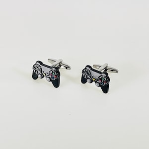 May include: A pair of cufflinks shaped like video game controllers. The cufflinks are black with silver-colored metal accents. The controller buttons are detailed with various colors, including red, green, and blue.