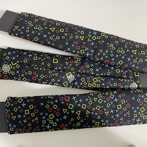 May include: Three black fabric seatbelt covers with a colorful geometric pattern of squares, circles, triangles, and crosses. The pattern is in shades of yellow, red, blue, and green.