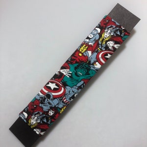 Puede incluir: Un objeto rectangular cubierto de tela, que muestra un collage de personajes de cómics de Marvel, incluyendo a Spider-Man, Capitán América y Hulk. La tela está envuelta alrededor de una base rectangular gris oscuro. El diseño es una vibrante mezcla de rojo, azul, verde y amarillo.