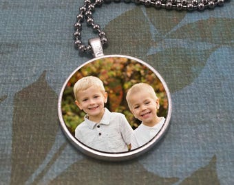 May include: A round silver pendant necklace featuring a photograph of two young boys smiling. The pendant is attached to a silver-toned ball chain. The image inside the pendant shows the boys against a blurred background of autumn foliage.
