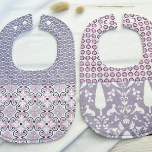 Peut inclure: Deux bavoirs pour bébé à motifs violets et blancs avec des fermetures à pression. Les bavoirs ont des motifs différents, l'un avec un motif géométrique et l'autre avec un motif floral.