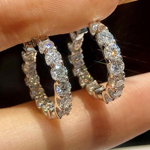 以下が含まれることがあります： 銀色のフープピアス一対。金属製のフレームに、透明で丸いファセットカットの石が一列に並んでセットされています。石は高い輝きを放ち、光を反射してキラキラと輝く多面的な表面を持っています。