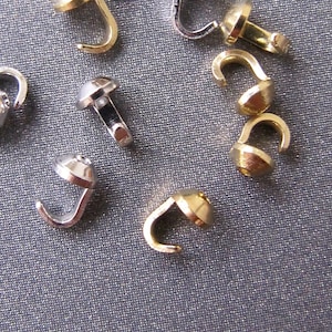 Op de afbeelding: Een verzameling kleine, gouden en zilveren metalen haken met een afgeronde bovenkant. De haken zijn in een cirkel op een grijs oppervlak gerangschikt.