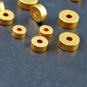 Könnte beinhalten: Eine Sammlung goldfarbener Metallperlen mit einem Loch in der Mitte. Die Perlen sind in verschiedenen Größen und auf einer dunkelgrauen Oberfläche verstreut.