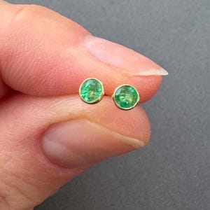 Puede incluir: Un par de pequeños pendientes de botón redondos de esmeralda con engastes de oro. Las piedras preciosas de color verde brillante están engastadas en un diseño sencillo y clásico. Los pendientes se muestran sobre un fondo neutro.