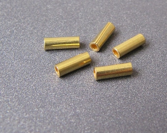 May include: Five small, cylindrical brass tubes with a polished finish are scattered on a gray surface. Each tube has a hollow center and is approximately 0.5 inches long. The tubes are evenly spaced and appear to be components for jewelry making or other crafts.