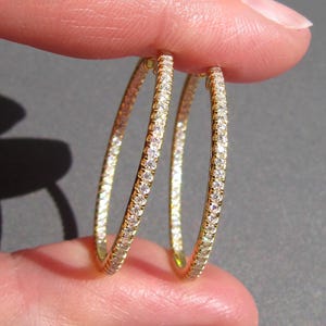 May include: A pair of gold hoop earrings featuring a continuous row of small, clear, round-cut stones set along the outer edge of each circular band. The earrings have a polished metallic finish and a thin, lightweight, circular design.
