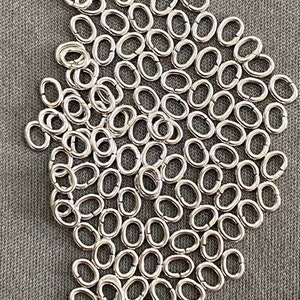 May include: A large pile of silver jump rings, a type of jewellery finding, on a grey fabric background.