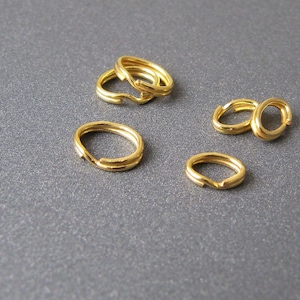 Puede incluir: Cinco anillos de salto dorados, también conocidos como anillos partidos, están dispuestos sobre una superficie gris. Los anillos son de diferentes tamaños y se utilizan comúnmente en la fabricación de joyas.