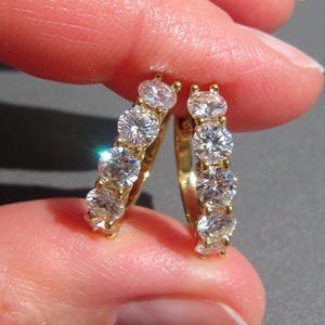May include: A pair of gold hoop earrings, each featuring a row of four round, clear, faceted stones set in a vertical line. The metal is a warm yellow tone, and the stones exhibit high clarity and brilliance, reflecting light from the surface.
