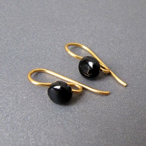 Puede incluir: Un par de pendientes de gancho dorados, cada uno con una pequeña cuenta negra redonda y brillante. Los pendientes se muestran sobre un fondo gris, resaltando su diseño simple pero elegante. Los ganchos son curvos y delicados.