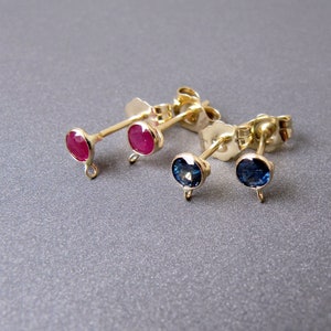 Puede incluir: Tres pares de pendientes de botón de oro con pequeñas piedras redondas. Un par tiene piedras rojas y los otros dos pares tienen piedras azules.