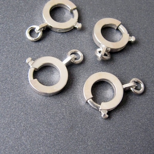 Puede incluir: Cuatro cierres de metal plateados para la elaboración de joyas. Cada cierre es un círculo con una abertura de resorte.