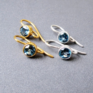 Op de afbeelding: Een paar oorbellen met blauwe topaas edelstenen. Het ene paar is goud en het andere is zilver.