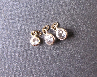 May include: Three gold-colored pendants with clear, faceted gemstones. Each pendant has a small loop at the top for attaching to a chain or bracelet. The gemstones are oval and round.