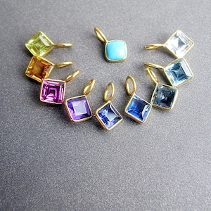 Puede incluir: Un conjunto de diez dijes de oro con piedras preciosas cuadradas de diferentes colores. Los dijes están dispuestos en semicírculo con un dije turquesa en el centro.