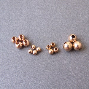 May include: A collection of small, round, gold-coloured beads of varying sizes. The beads are arranged in three groups, with the largest group in the centre.