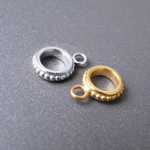 May include: Two small, round metal charms. One is silver-colored, the other is gold-colored. Both charms have a textured, beaded design around the outer edge and a small loop for attaching to jewelry or other crafts. The background is a solid gray.