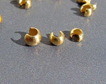 May include: A collection of small, gold-colored crimp beads scattered on a gray surface. The beads have a C-shaped design with a small opening, and are likely used in jewelry making or crafting projects. The lighting highlights the metallic sheen of the beads.