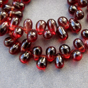 May include: A strand of deep red gemstone beads in a teardrop shape. The beads are strung on a clear thread and are arranged in a circular pattern.