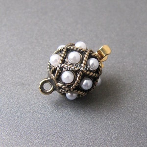 Könnte beinhalten: Ein runder, goldfarbener Verschluss mit perlengesetztem Design. Der Verschluss ist aus Metall und hat eine kleine Schlaufe an der Seite.