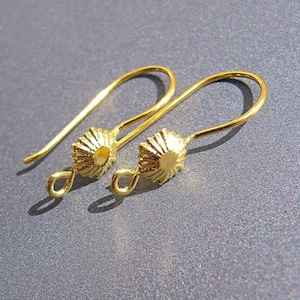 May include: A pair of gold-colored earrings. Each earring features a decorative, sunburst-shaped bead and a curved ear wire. The earrings are displayed on a gray surface, with shadows adding depth. The jewelry is designed for pierced ears.