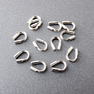 Puede incluir: Primer plano de terminales de crimpado de metal plateado para la elaboración de joyas. Las terminales tienen forma de U y están dispuestas de forma dispersa sobre una superficie gris.