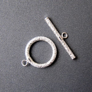 May include: Silver toggle clasp with a textured finish. The clasp has a round ring and a straight bar.