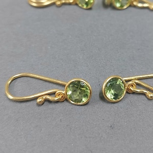 Könnte beinhalten: Ein Paar goldene Ohrringe mit grünen Edelstein-Akzenten. Die Ohrringe haben ein schlichtes, elegantes Design mit einem kleinen, runden Edelstein in der Mitte jedes Ohrrings.