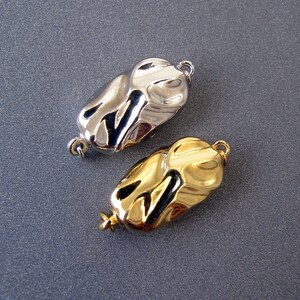 Op de afbeelding: Twee metalen sluitingen voor het maken van sieraden. E&eacute;n is zilver en de andere is goud. Beide hebben een textuur en een organische vorm.