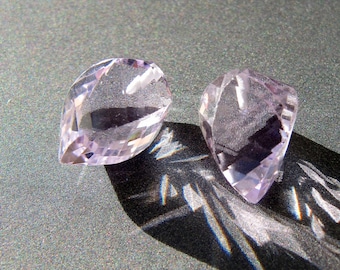 May include: Two faceted, pear-shaped gemstones in a soft lavender hue. The gems are positioned on a textured, dark gray surface, casting a sharp shadow. The light catches the facets, creating a sparkling effect.