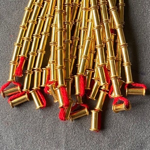 Könnte beinhalten: Nahaufnahme von goldfarbenen Metallperlen, die mit rotem Faden aufgefädelt sind. Die Perlen haben eine zylindrische Form mit einem segmentierten Design. Der rote Faden ist an den Enden der Stränge sichtbar und bildet einen Kontrast zum Gold.