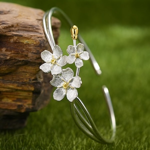 Könnte beinhalten: Silbernes Armreif-Armband mit drei zarten weißen Blumenanhängern und einem goldenen Akzent. Das Armband ist offen und verstellbar.