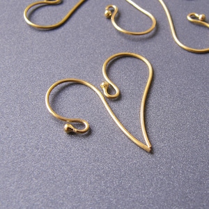 Puede incluir: Primer plano de varios ganchos de pendientes dorados, algunos en forma de corazón. Los ganchos están hechos de alambre fino y tienen un diseño simple y elegante. El fondo es de color gris oscuro sólido.