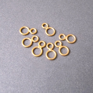 May include: Gold-toned metal jump rings with a double loop design. The rings are arranged in a semi-circle on a gray surface.