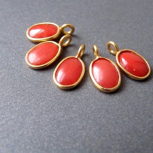 以下が含まれることがあります： 鮮やかな赤色の中心と金色のフレームが付いた、5つの楕円形のペンダント。各ペンダントの上部には、チェーンや他のジュエリーコンポーネントに取り付けるための小さなループが付いています。ペンダントは灰色の表面に表示されています。