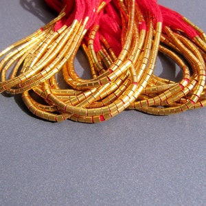 Puede incluir: Un montón de cuentas doradas con hilo rojo tejido a través de ellas. Las cuentas son pequeñas y redondas, y están dispuestas en un patrón suelto y fluido.