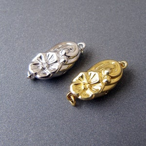 May include: Two ornate metal clasps, one silver and one gold, shaped like a flower with a rounded oval base.