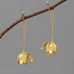 Puede incluir: Un par de pendientes colgantes de color dorado con un delicado diseño floral. Los pendientes están suspendidos de un alambre fino y tienen un estilo simple y elegante.