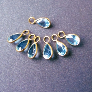 May include: A collection of teardrop-shaped blue gemstones with gold-colored metal settings. The gemstones are arranged on a dark blue surface, showcasing their clarity and the contrast with the gold. These are likely jewelry components.