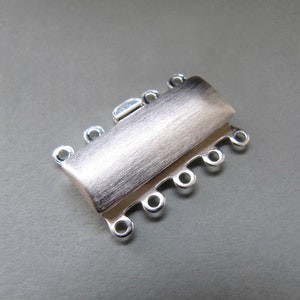 Puede incluir: Un cierre de joyería plateado de múltiples hilos. El cierre tiene un cuerpo rectangular con un acabado de metal cepillado y múltiples bucles para sujetar los hilos. El cierre está diseñado para asegurar múltiples hilos de un collar o pulsera.