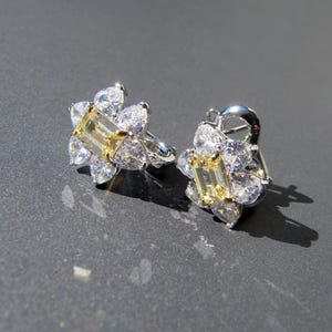 Pode incluir: Um par de brincos de botão em forma de flor. Cada brinco apresenta uma gema amarela retangular central rodeada por pedras brancas em forma de pêra. Os brincos são cravejados em metal prateado.