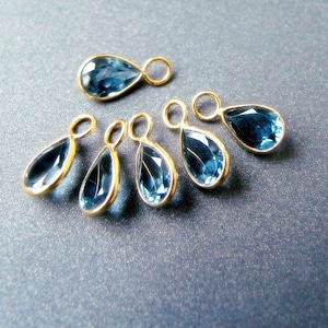 Op de afbeelding: Zes traanvormige blauwe edelsteenbedels met goudkleurige springringen. De bedels zijn in een halve cirkel op een blauwe achtergrond gerangschikt.