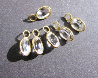 May include: Several gold-colored oval pendants with clear, faceted stones. Each pendant has a small loop at the top for attaching to a chain or earring hook. The pendants are displayed on a dark gray surface.