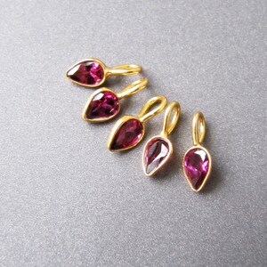 Op de afbeelding: Vijf gouden traanvormige bedels met roze edelstenen. De bedels hebben een lus aan de bovenkant om aan sieraden te bevestigen.